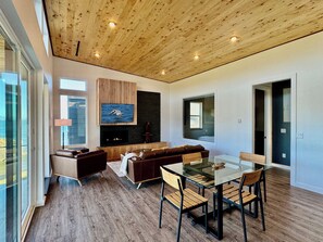 A modern living room with a glass dining table, wooden chairs, a sofa, and a fireplace.