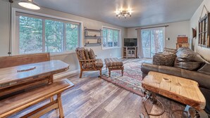 A spacious living room with a wooden dining table, a patterned armchair, a leather sofa, and a large window providing a view of the outdoors.