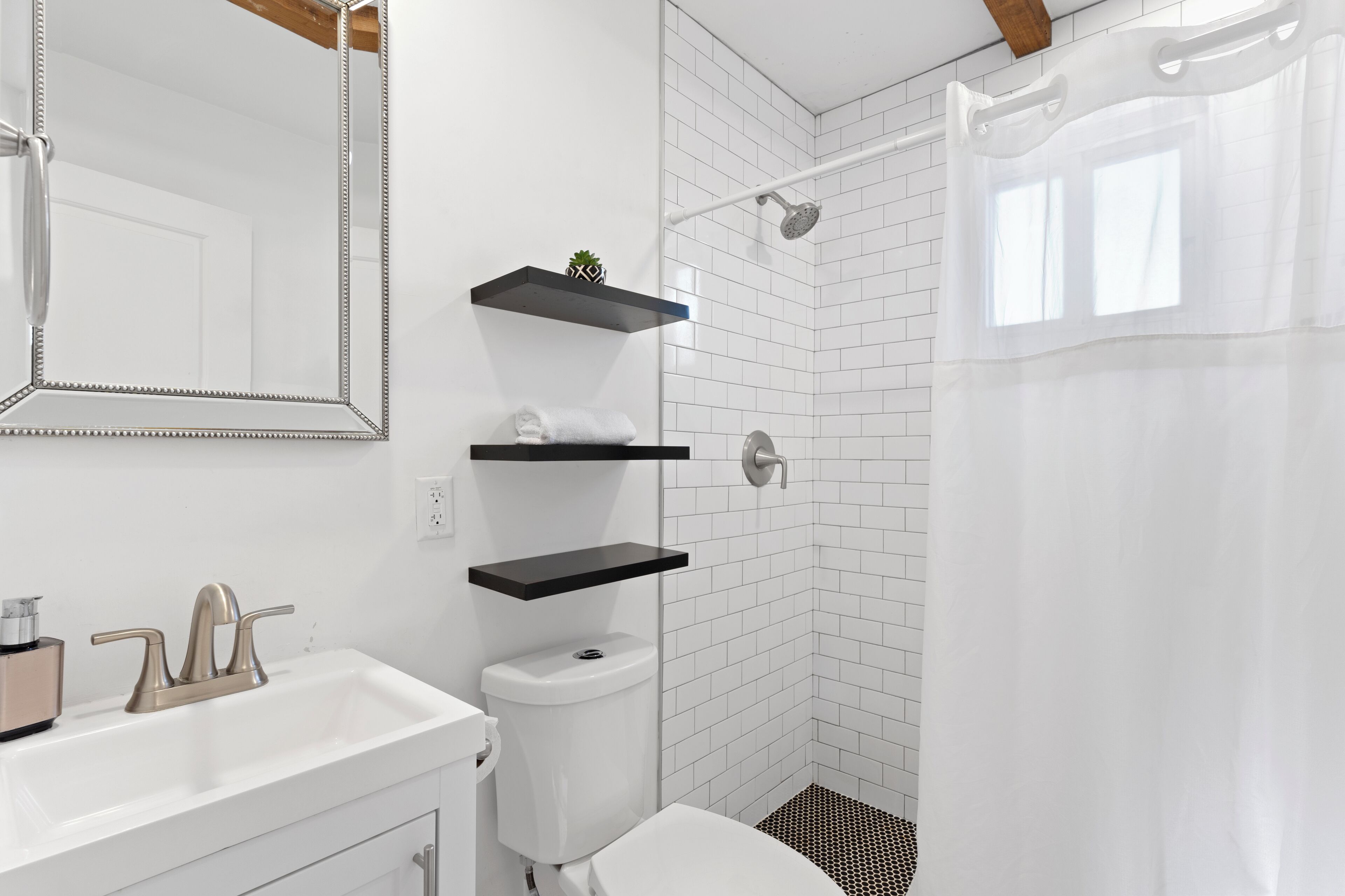 Une salle de bain moderne avec un lavabo blanc, des toilettes, une douche avec une porte en verre et deux étagères noires fixées au mur.
