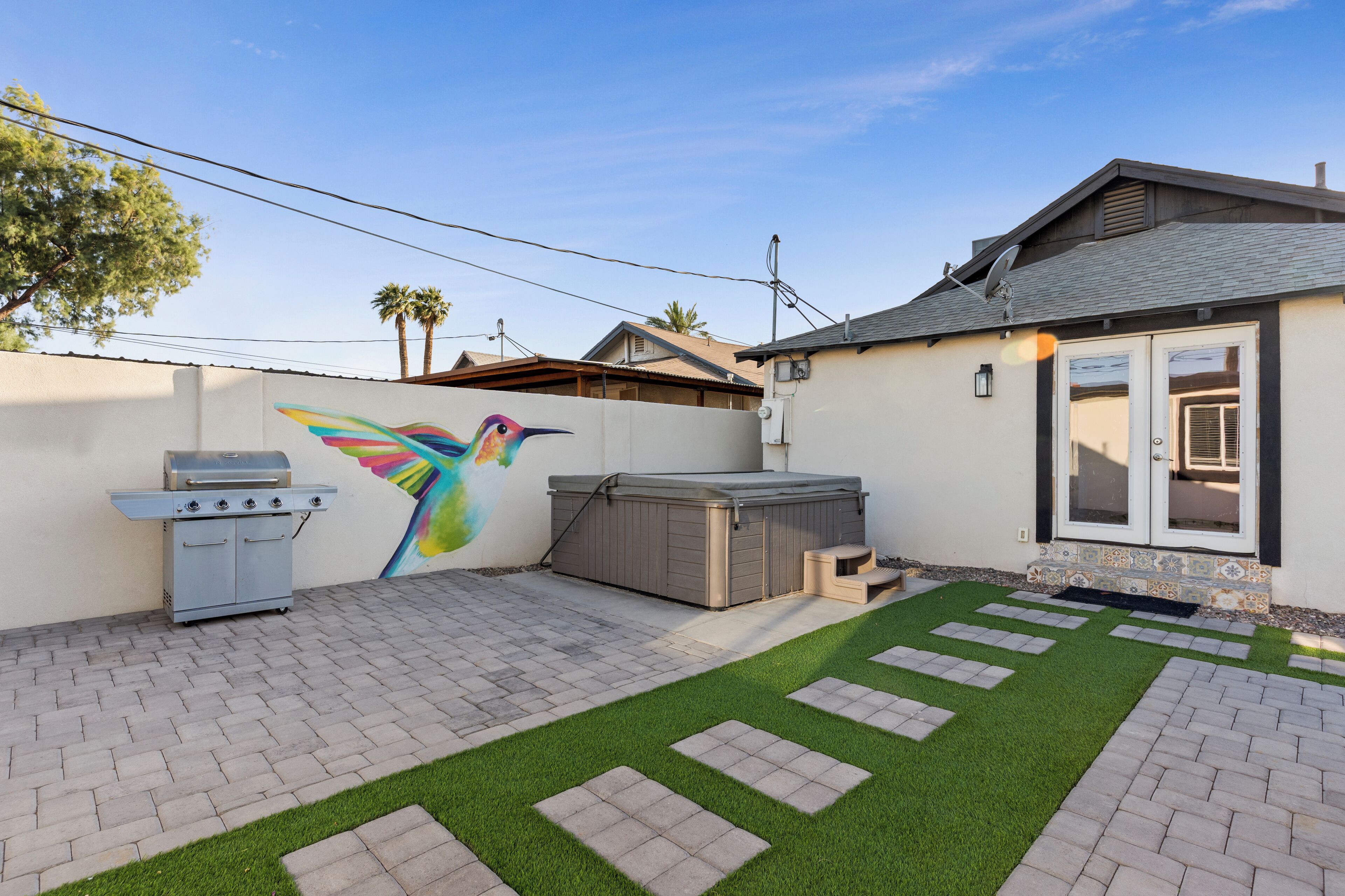 A backyard with artificial grass, a barbecue grill, a hot tub, and a colorful mural of a hummingbird.