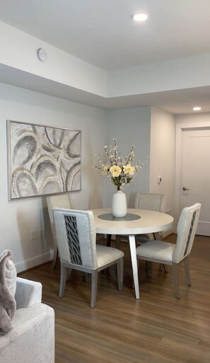 Un comedor con una mesa redonda, cuatro sillas, un florero con flores, una obra de arte colgada en la pared y una puerta al fondo.
