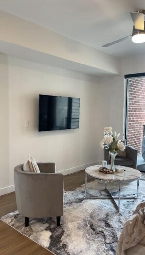 A modern living room with a flat-screen TV mounted on the wall, a grey armchair, a round coffee table with a vase of flowers, and a large window with a brick wall view. - Buckhead Atlanta Luxury Suite (Atlanta)
