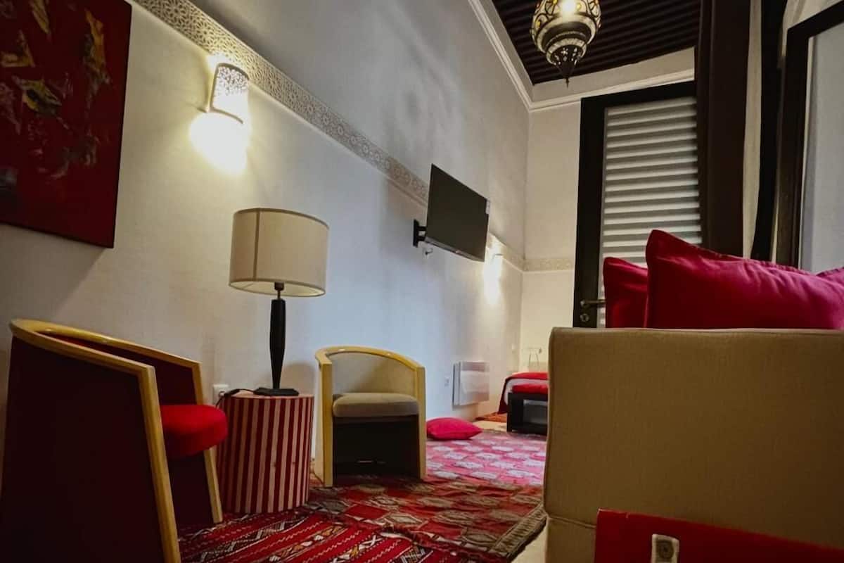 A hotel room with a patterned carpet, a bed with red pillows, a sofa, a small table with a lamp, and a TV mounted on the wall.