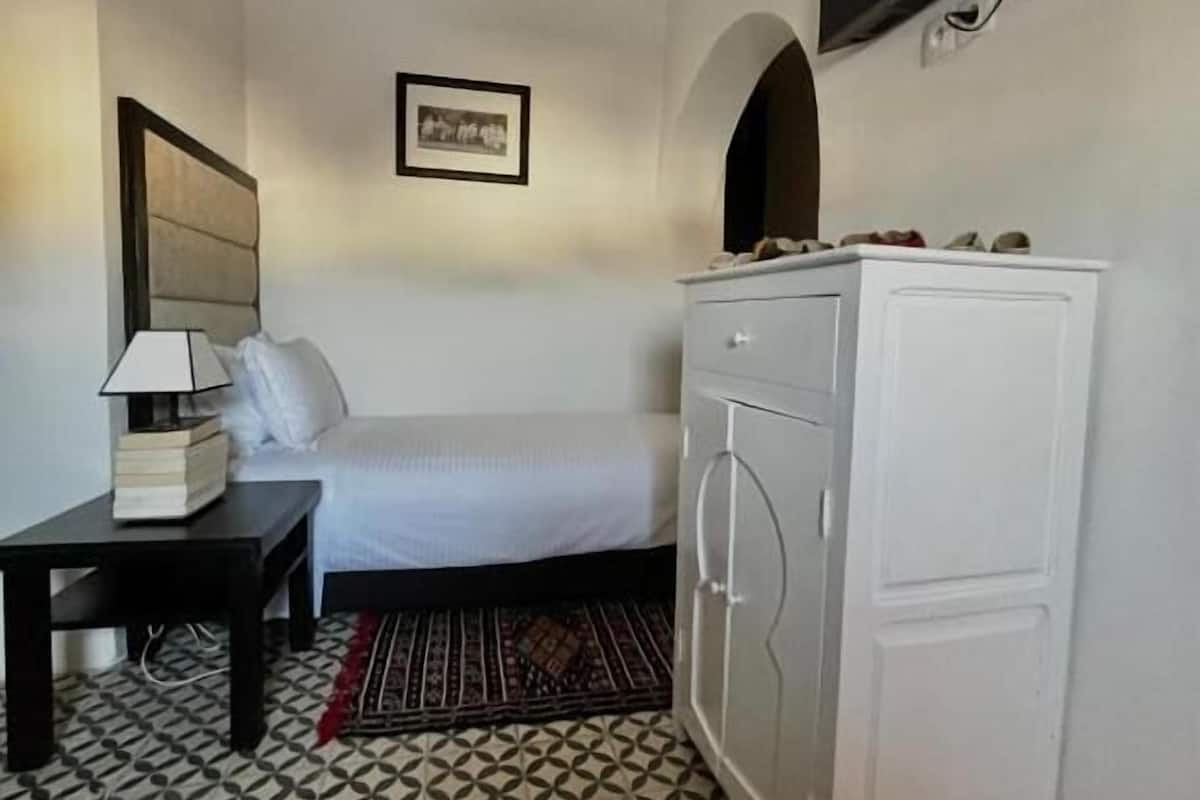 A hotel room with a bed, a bedside table with a lamp, a white wardrobe, and a patterned rug.