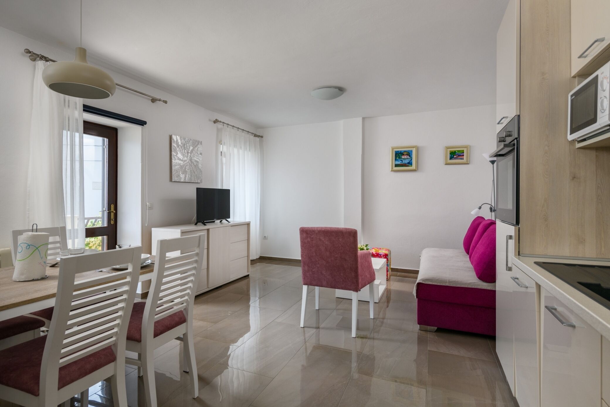Una cocina moderna con armarios blancos, un comedor con sillas blancas, un sofá rosa y un televisor.