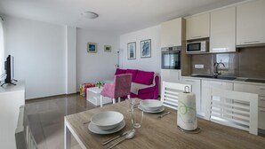 A modern kitchen with white cabinets, a microwave, and a sink. There is a dining table with plates, cutlery, and a glass. A pink chair is visible, and a sofa is in the background. - Apartment Yasmin (Kvarner - Mošćenička Draga) (Mošćenička Draga)