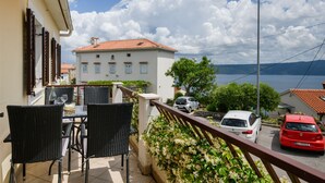 A balcony with wicker chairs and a table, overlooking a street with parked cars and a building with a red roof. - Apartment Yasmin (Kvarner - Mošćenička Draga) (Mošćenička Draga)