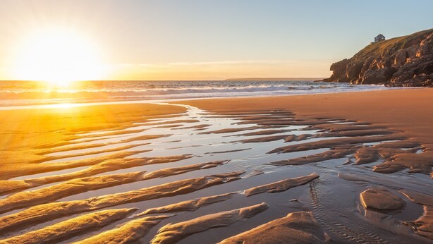 Una playa con atardecer, olas y un acantilado rocoso con una estructura en la cima.