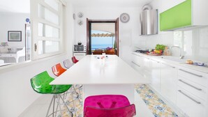 A modern kitchen with a view to the sea, white cabinets, and colorful chairs. - Villa Mapi (Massa Lubrense)