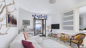 A modern living room with a white sofa, a wooden chair, a coffee table, and a TV. There is a balcony with a view of the sea. - Villa Mapi (Massa Lubrense)