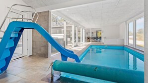 Una piscina interior con un tobogán azul, puertas de cristal que dan a un área exterior y vistas al exterior.