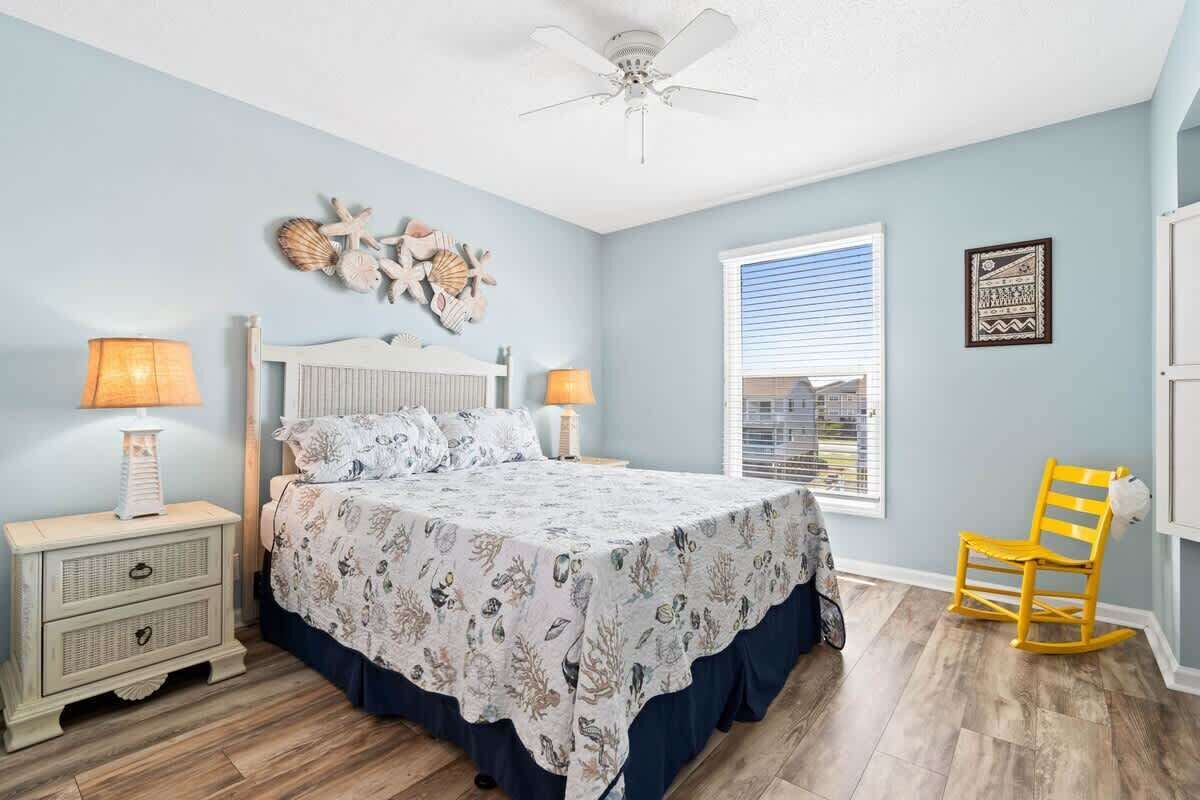 A bedroom with a bed, two bedside tables, a ceiling fan, a window with blinds, a chair, and wall decorations.