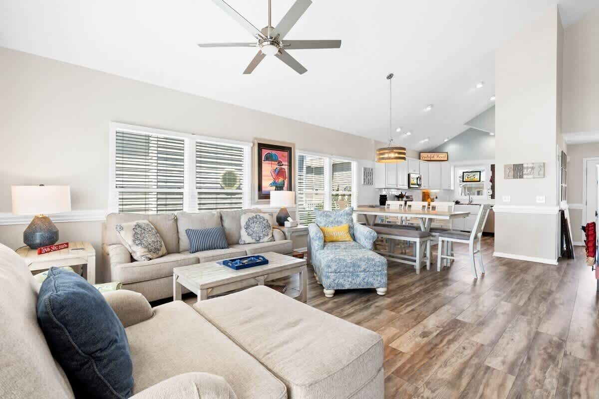 A spacious living room with a sofa, armchair, and coffee table, a dining area with a table and chairs, and a kitchen with white cabinets and a ceiling fan.