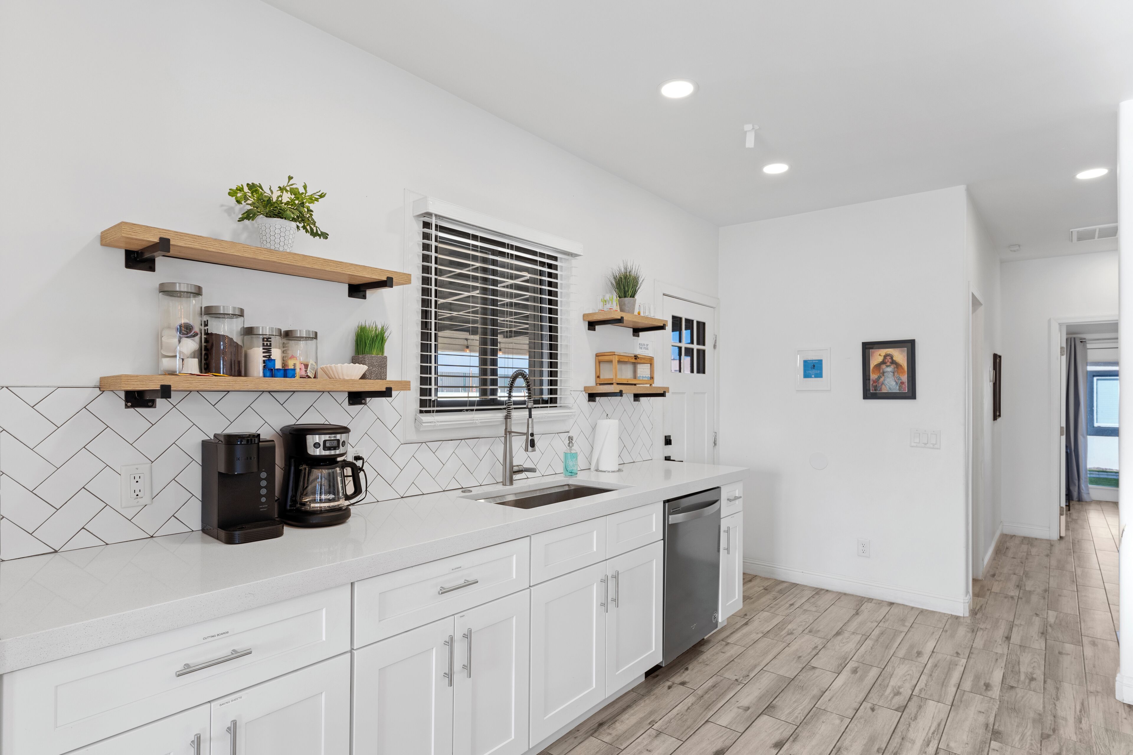 A modern kitchen with white cabinets, a built-in dishwasher, a coffee maker, and a window with blinds.