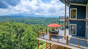 Una terraza de madera de dos pisos con un paraguas rojo, mobiliario exterior y vistas a un bosque pintoresco.