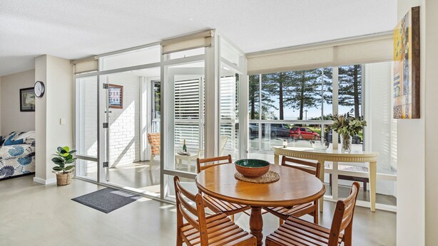 A dining area with a round wooden table and chairs, a view of trees and cars through large windows, and a white brick wall. - Rainbow Pacific Ocean Views (Coolangatta)