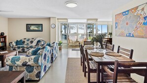 A spacious living room with a sofa, a dining table with chairs, a bookshelf, and a large window with blinds. - Rainbow Pacific Ocean Views (Coolangatta)