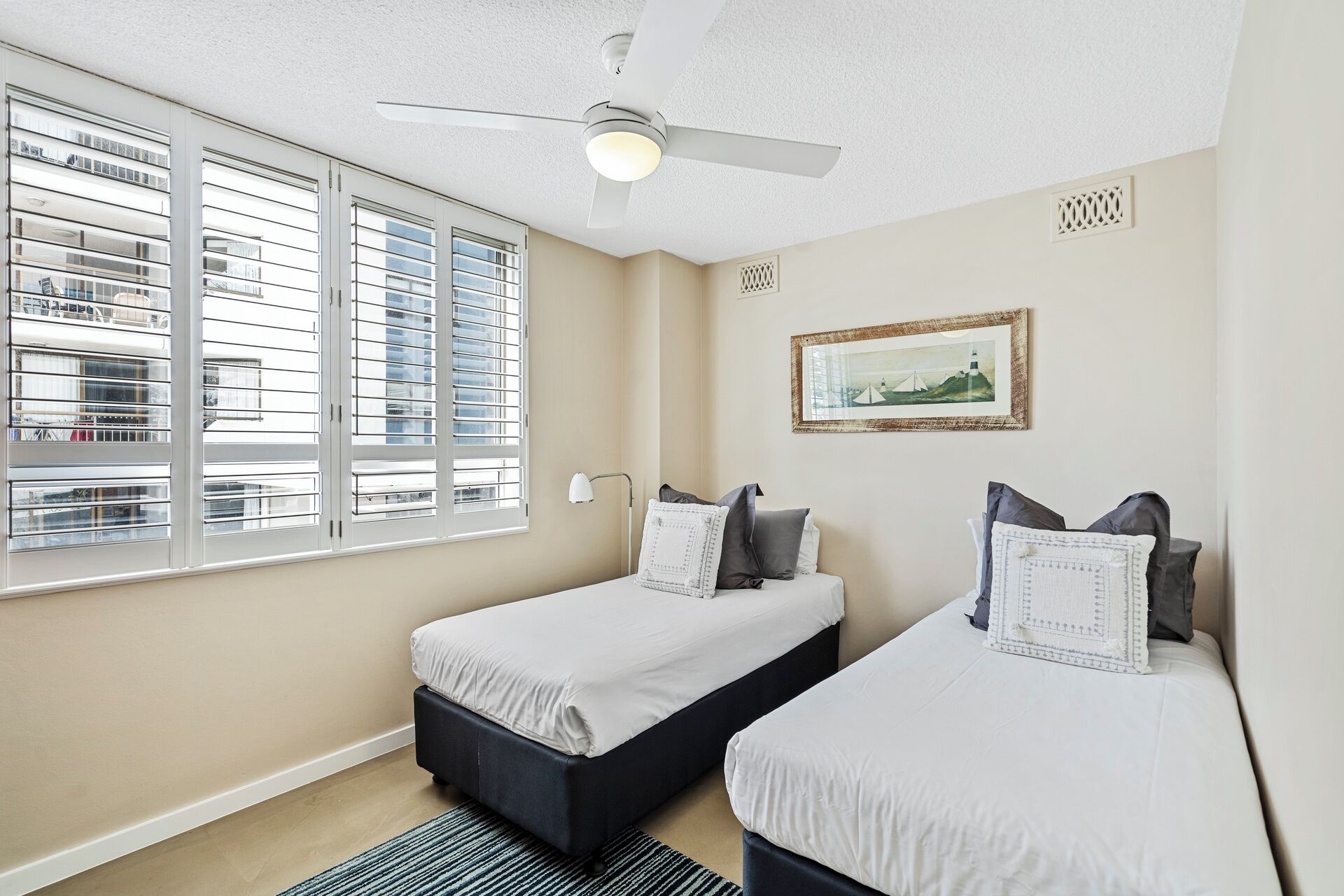 A bedroom with two beds, a ceiling fan, a window with blinds, and a framed picture on the wall.