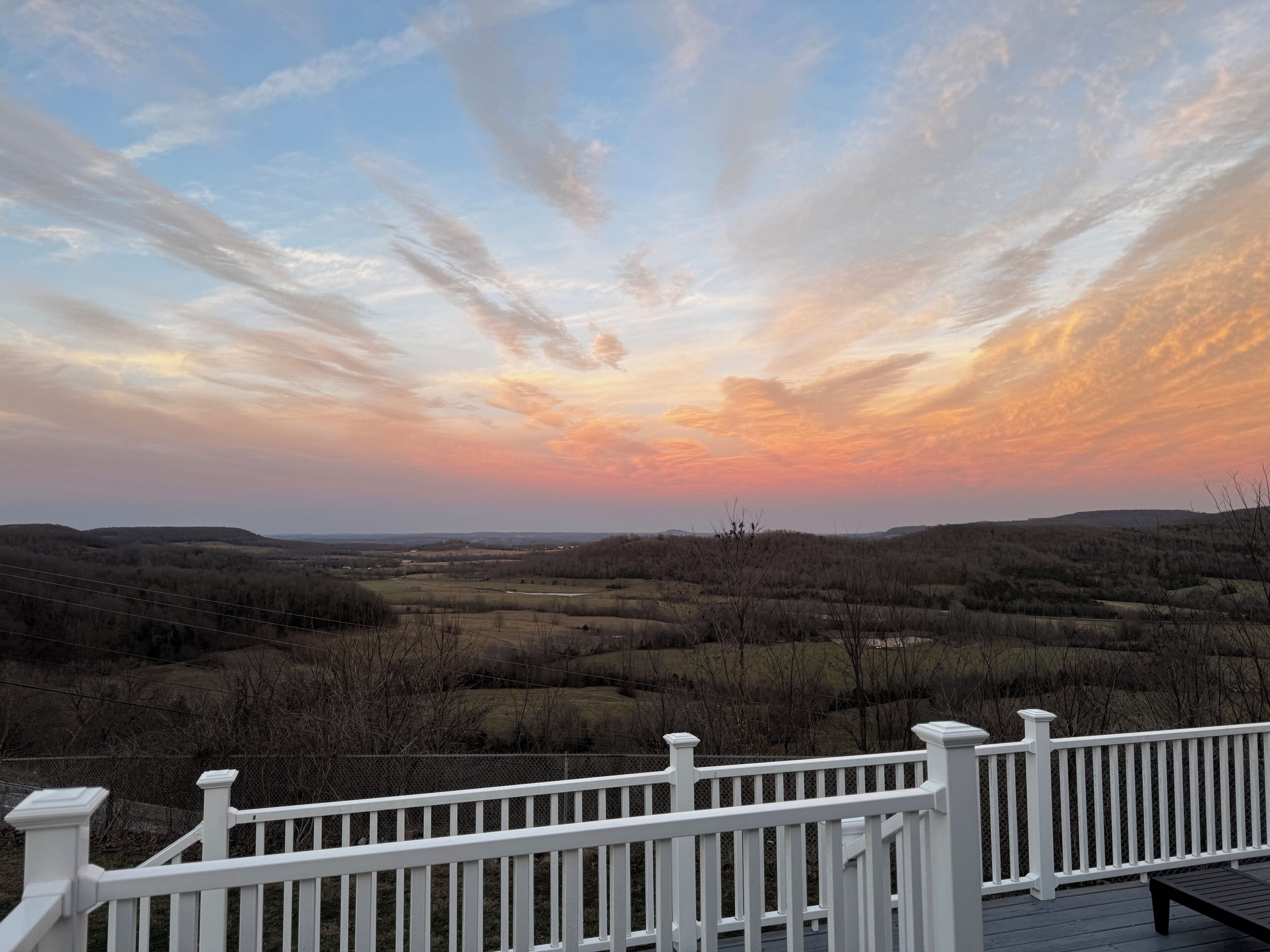 Eine Terrasse mit weißer Reling, die auf eine Landschaft mit sanften Hügeln und einem Sonnenuntergangs-Himmel blickt.