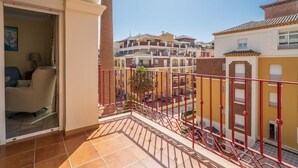Un balcón con barandilla roja, vista a edificios vecinos y una puerta que da a una habitación con una silla y una lámpara.