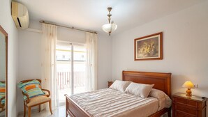 Un dormitorio con cama de madera, una mesita de noche con lámpara, una silla con cojín, un cuadro en la pared y un balcón con cortinas.