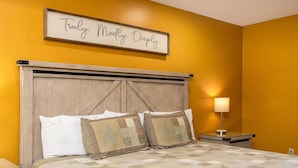 A bedroom with a bed, two decorative pillows, a nightstand with a lamp, and a sign above the bed.