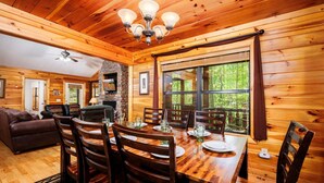 Interior de una cabaña de madera con una mesa de comedor preparada para cuatro personas, chimenea y ventanas amplias.