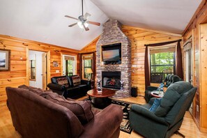 L’intérieur d’une cabane en rondins, avec une cheminée en pierre, des murs en bois, un ventilateur de plafond et un téléviseur fixé au-dessus de la cheminée.