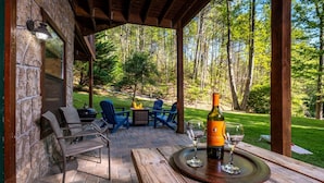 Un patio techado al aire libre con paredes de piedra, un hogar para fogata, sillas azules y una mesa con una botella y vasos.