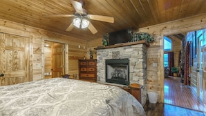 Une chambre située dans un chalet en rondins, avec un grand lit, une cheminée en pierre, un ventilateur de plafond et des murs en bois.