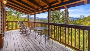 Une terrasse en bois avec une table et des chaises, offrant une vue sur un paysage boisé avec des montagnes à l’horizon.
