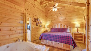 A wooden cabin bedroom with a bed, a nightstand, a lamp, and a bathroom with a bathtub.