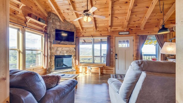 A cozy cabin interior with a stone fireplace, leather sofas, a flat-screen TV, and a balcony with a view of the mountains.