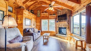 A cozy cabin interior with a stone fireplace, wooden furniture, and large windows offering a view of the outdoors.