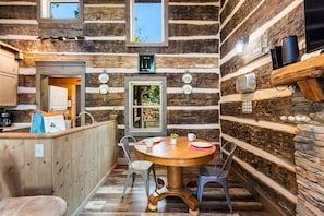 L’intérieur d’un chalet rustique comprend une table à manger en bois, des chaises, un coin cuisine et une grande fenêtre.