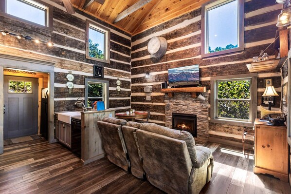 A log cabin interior with a fireplace, wooden walls, a kitchen area, and a seating area with a sofa.