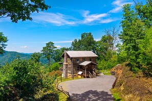 A wooden cabin with a porch, surrounded by lush greenery and a clear sky.
