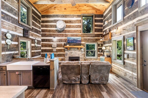 L’intérieur d’une cabane en rondins est doté d’une cheminée en pierre, de murs en bois, d’un coin cuisine et d’un espace salon avec un téléviseur.
