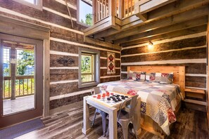 Une chambre située dans un chalet en rondins, avec un lit, une table sur laquelle est posé un jeu de société et une fenêtre donnant sur la verdure.