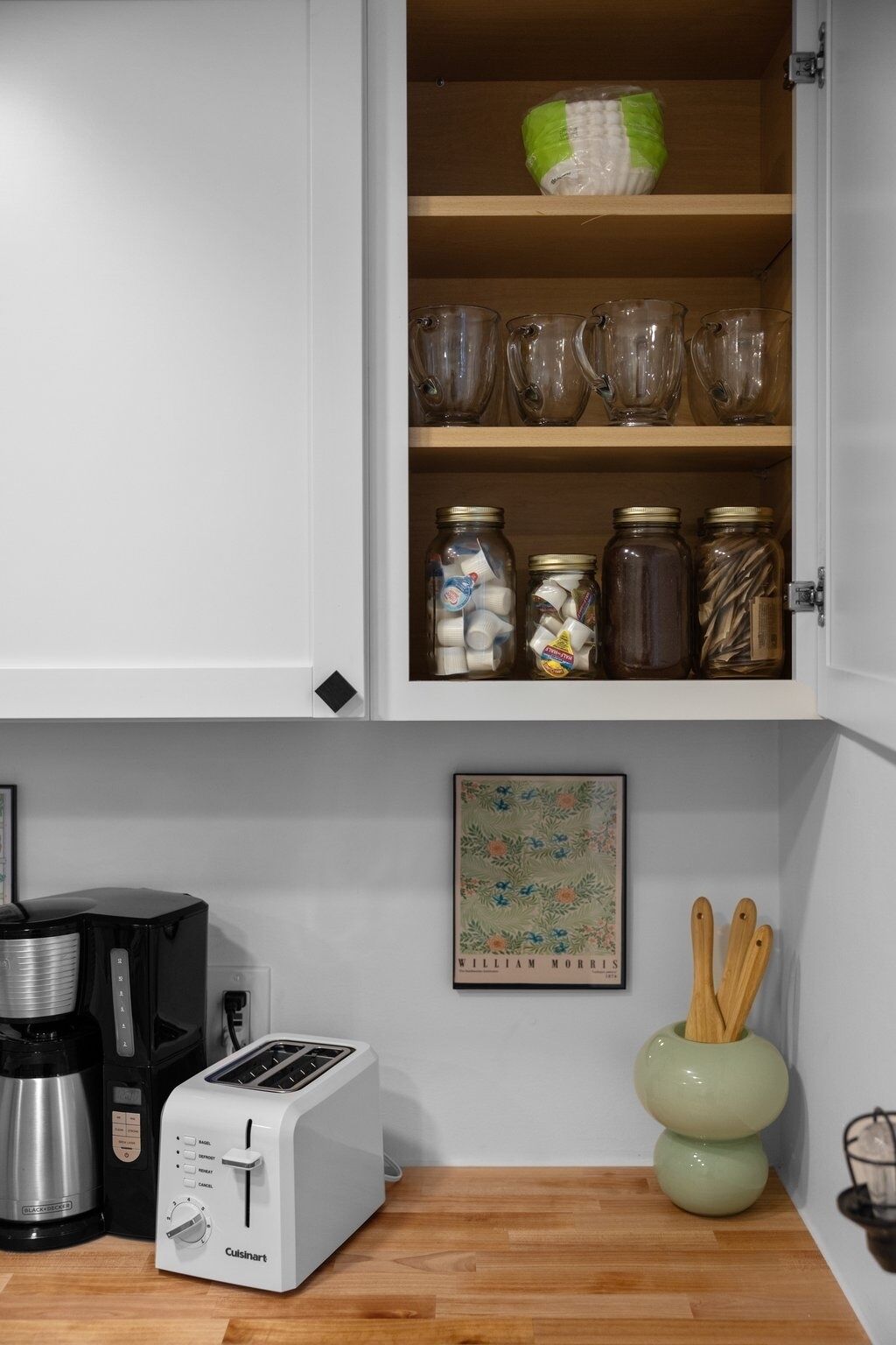 A kitchen with a white cabinet containing glassware and jars, a Cuisinart toaster, a coffee maker, and a framed botanical print.