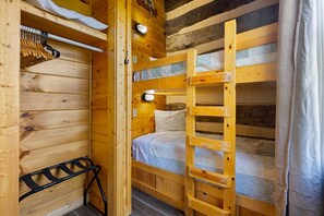Habitación con literas de madera, una escalera, un perchero y una lámpara fijada a la pared.