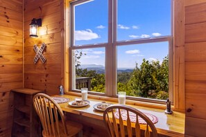 Interior de una cabaña de madera con una mesa de comedor, sillas, una ventana con vistas a las montañas y una señal de cruce en la pared.