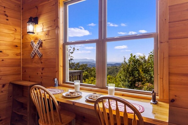 A wooden cabin interior with a dining table, chairs, a window with a view of the mountains, and a crossroad sign on the wall.
