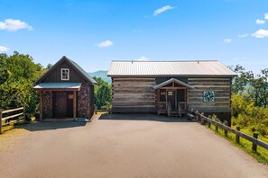 A rustic log cabin with a stone foundation and a metal roof, a wooden structure with a metal roof, and a paved driveway leading to a wooden fence. - The Homestead & The Shed - 2 side-by-side cabins! (Gatlinburg)