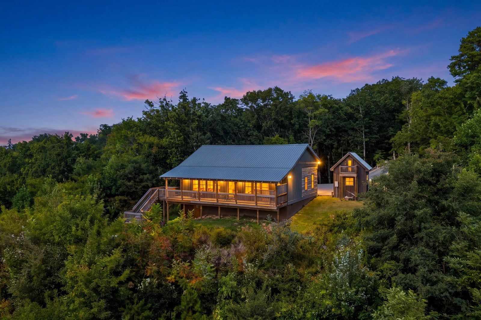 A cabin with a covered deck and a separate building, surrounded by dense forest.
