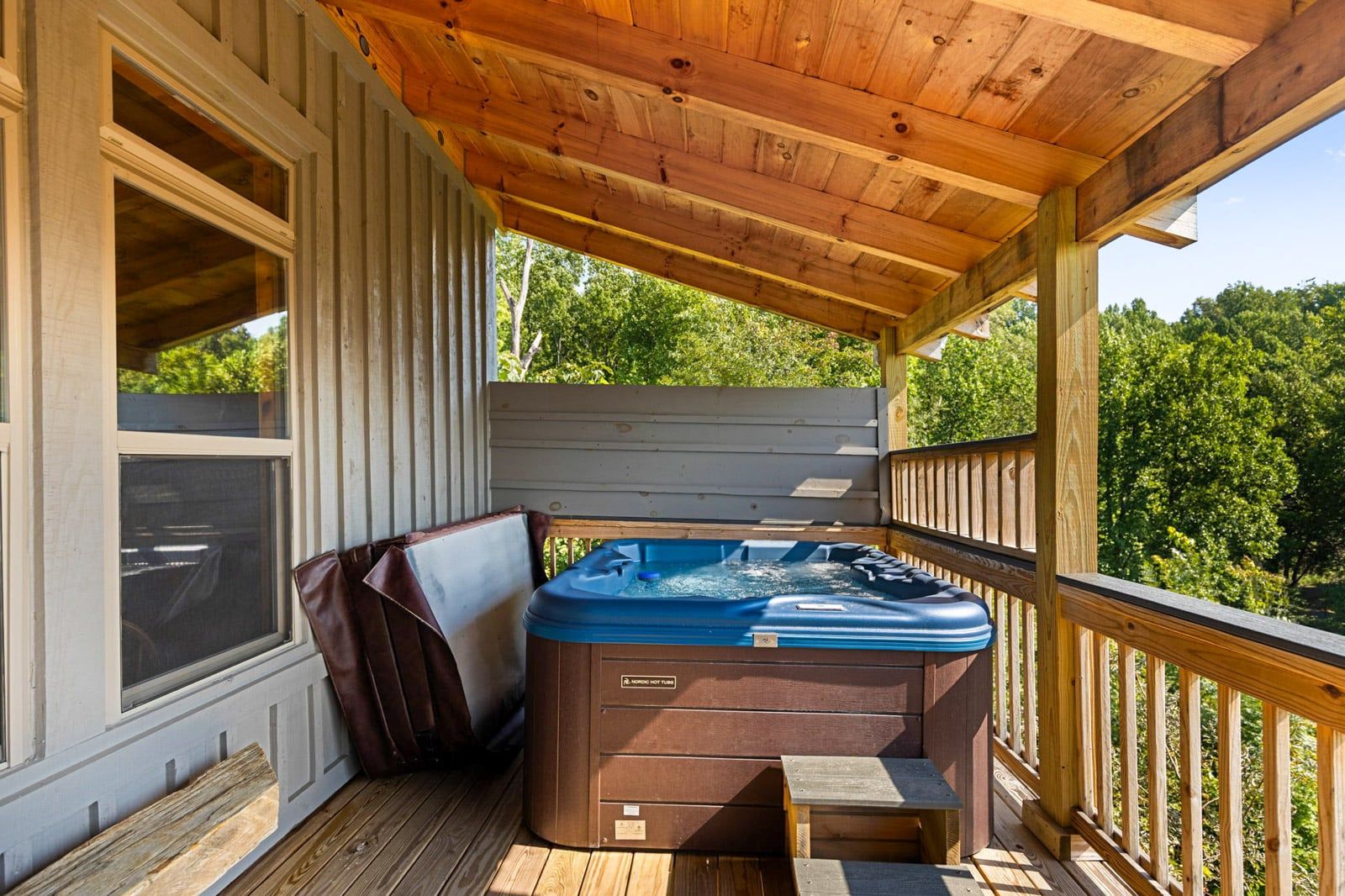 A wooden deck with a hot tub, a bench, and a view of greenery.