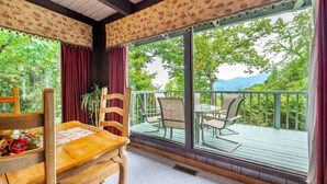 Una terraza de madera con una mesa, sillas, una planta en maceta y vistas a árboles y montañas a través de amplios ventanales.