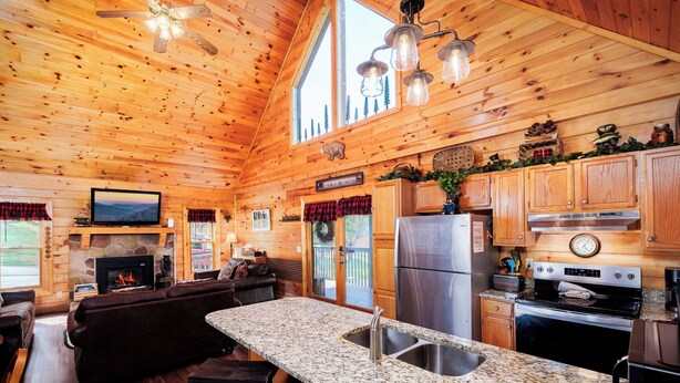 A log cabin interior with a stone fireplace, wooden furniture, granite countertops, and a kitchen with stainless steel appliances.