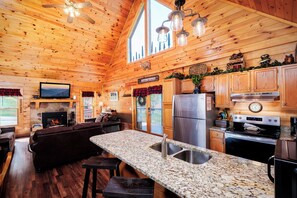 L’intérieur d’une cabane en rondins est agrémenté d’une cheminée en pierre, de meubles en bois, de plans de travail en granit et d’une cuisine équipée d’appareils électroménagers en acier inoxydable.