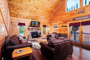 A log cabin interior with a fireplace, leather couches, a television, and a sign reading 'Great Smoky Mountains.' - Bear Necessities – Hot Tub, Game Loft & Lone Branch Creek Resort (Sevierville)